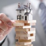 A close-up shot of a hand pulling out a Jenga block with a model house sitting on top of the tower.. Home prices. housing market layoffs