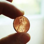 Image of a penny held between two fingers with a white indoor background